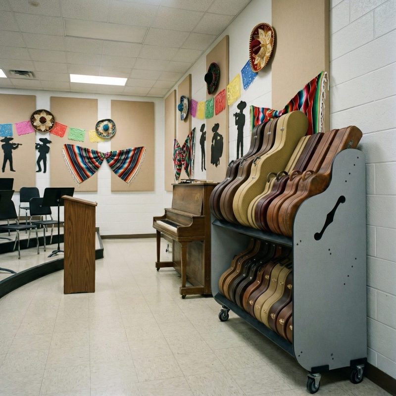 Two-tiered cart holding cases for mariachi instruments, next o a piano in a hispanic-themed classroom